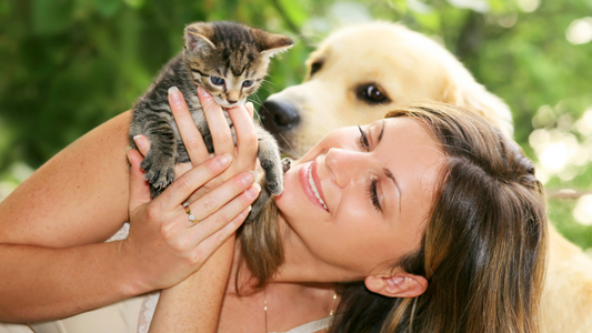 Smiling woman holding a small tabby kitten with a golden retriever looking on in the background, representing the emotional bond between female pet owners and their pets.